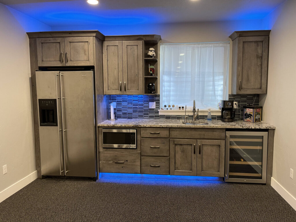 Modern basement kitchen with wood cabinets, granite countertop, wine fridge, microwave, and blue LED lighting