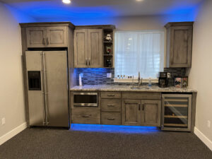 Modern basement kitchen with wood cabinets, granite countertop, wine fridge, microwave, and blue LED lighting